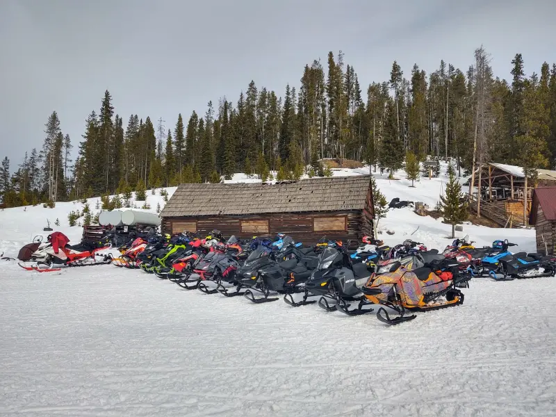 Dozens of snowmobiles parked in a long row outside a weathered log cabin with a cedar-shake roof, backed by conifers.