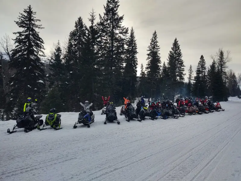A long line of about twenty-five snowmobiles and riders drawn up along a groomed road, several riders with an arm raised, beneath tall spruce.