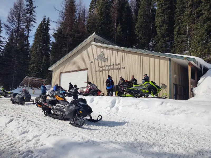 A metal club building with a snowmobile emblem and lettering across its gable, with snowmobiles and riders parked in front under a clear blue sky.