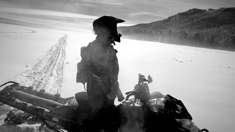 A black-and-white photograph of a helmeted rider standing on a snowmobile in close silhouette, with a long track running back across a wide frozen lake toward a timbered ridge.