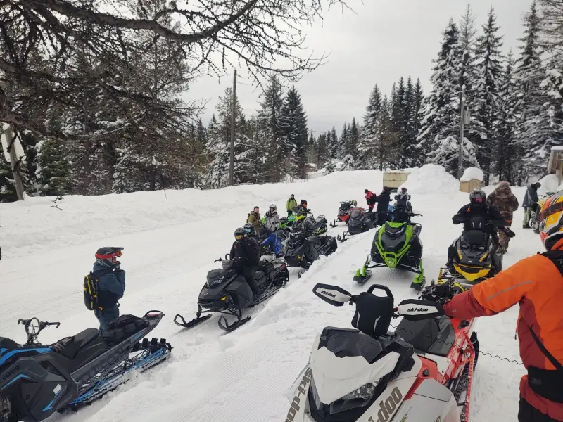Snowmobiles parked along a snow-packed forest road as riders in winter gear stand and talk beneath snow-laden spruce.