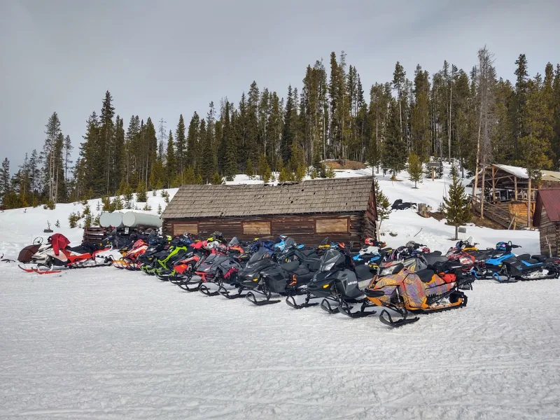 Dozens of snowmobiles parked in a long row outside a weathered log cabin with a cedar-shake roof, backed by conifers.
