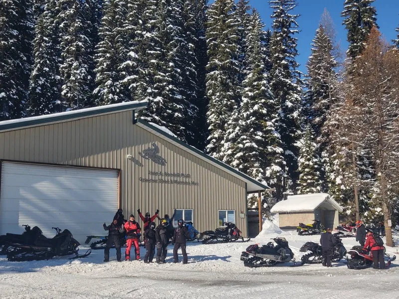 Riders and snowmobiles gathered outside a metal shop building with a snowmobile emblem on its gable, under a clear blue sky and heavily snow-laden spruce.