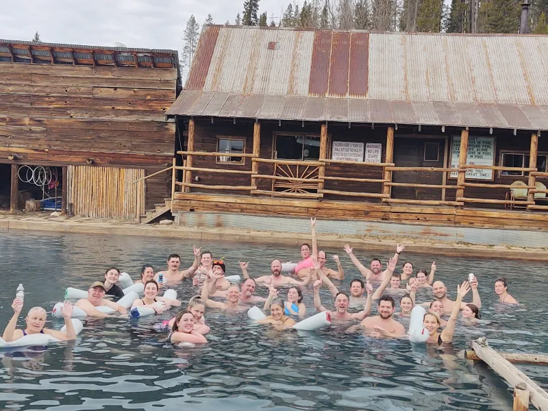 About thirty-five club members gathered in an outdoor hot spring pool, turned toward the camera, with rustic log bathhouse buildings behind them.