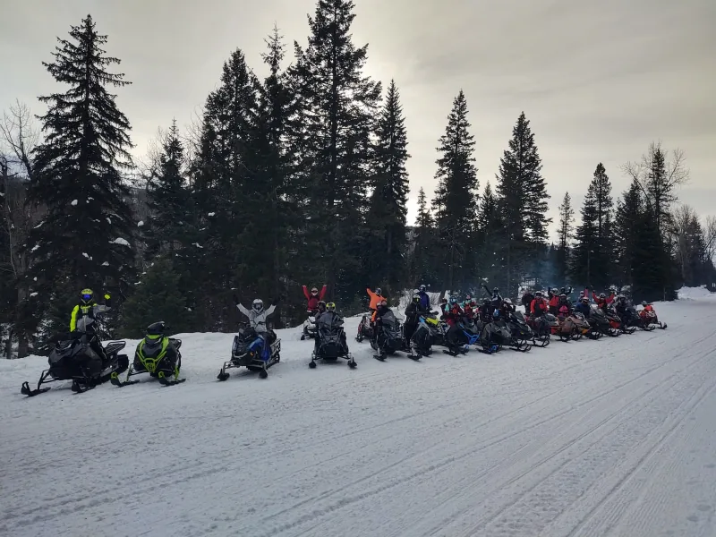 A long line of about twenty-five snowmobiles and riders drawn up along a groomed road, several riders with an arm raised, beneath tall spruce.