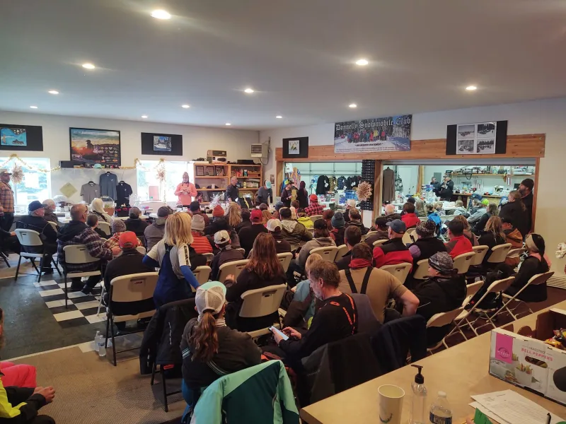 About eighty club members seated in folding chairs at an indoor meeting, facing a front table stacked with raffle baskets beneath a Donnelly Snowmobile Club banner.