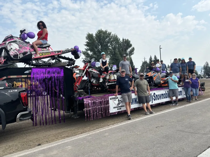 Club members standing beside a parade float — a flatbed trailer trimmed with purple streamers carrying snowmobiles and a snow bike, behind a banner reading Donnelly Idaho Snowmobile Club.