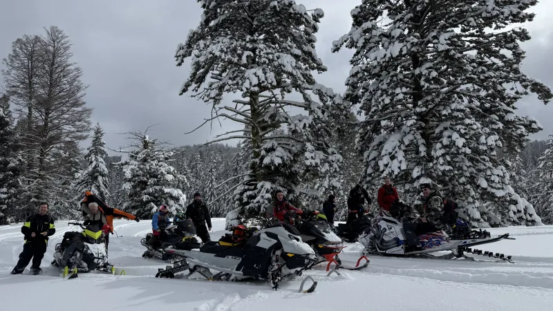 About ten riders standing and sitting with their snowmobiles in a snowy clearing beneath heavily snow-laden firs under an overcast sky.