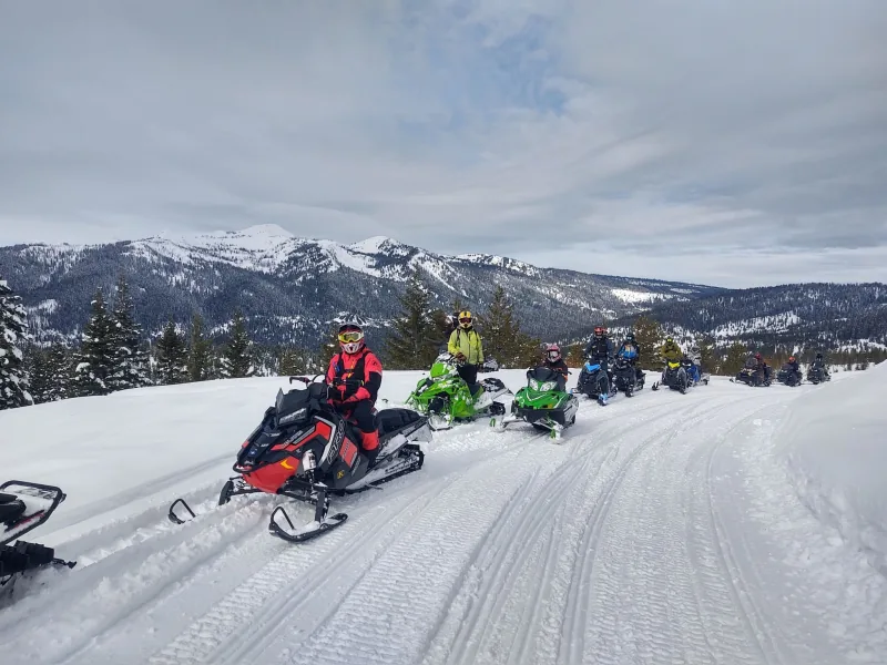 About ten riders stopped in a line along a groomed ridge road, with timbered slopes and a snow-covered peak behind them.