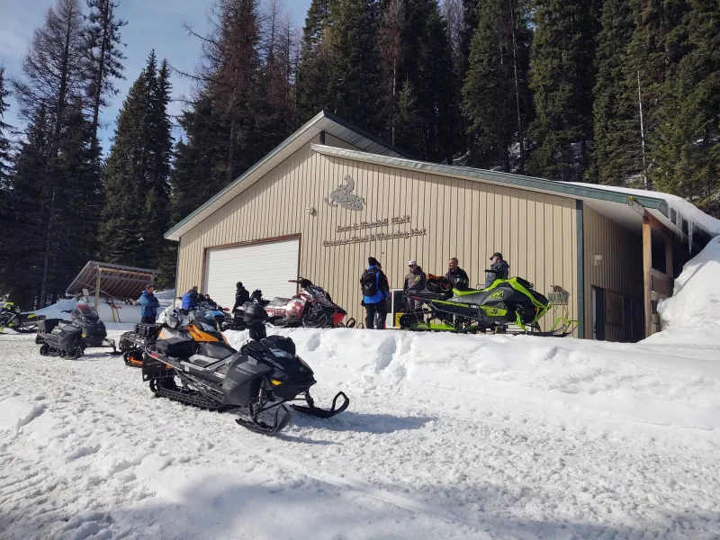 A metal club building with a snowmobile emblem and lettering across its gable, with snowmobiles and riders parked in front under a clear blue sky.