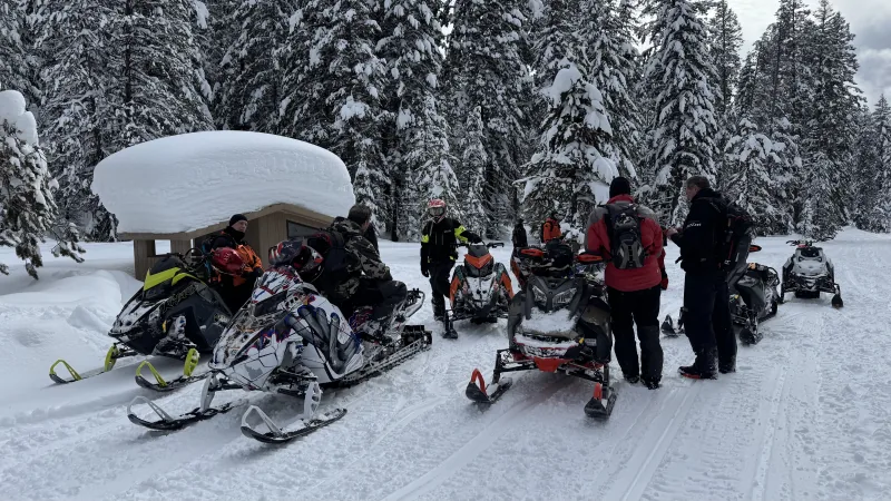 Riders and parked snowmobiles gathered beside a small trailside shelter capped with deep snow, among dense snow-covered conifers along a groomed road.